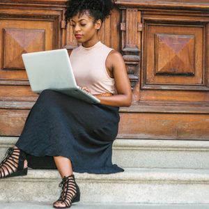 Person sitting cross-legged, using a laptop on stone steps, with wooden door backdrop.