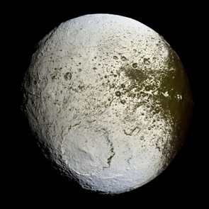 A heavily cratered, textured moon surface with contrasting light and dark regions, set against a black space background.