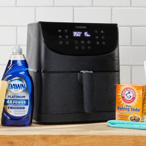Air fryer with illuminated display sits on a wooden counter, accompanied by a sponge, dish soap, baking soda, and toothbrush against a tiled backsplash.