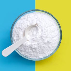 A glass bowl holds white powder with a white spoon resting inside, set against a background split between blue and yellow.