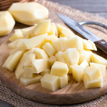 Chopped potatoes on a wooden board beside a knife; surrounding are whole, peeled potatoes on a woven mat surface.
