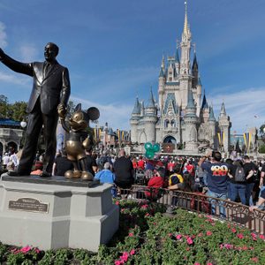 Statue features a man and mouse standing, with a castle and large crowd in the background, surrounded by vibrant flowers and clear skies.