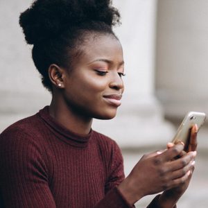 Woman holding a smartphone, smiling slightly, seated outdoors with blurred architectural background.