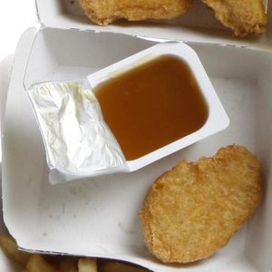 Chicken nuggets and a sauce container sit in a white cardboard box, surrounded by French fries and a burger on the side.