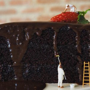 Miniature figures paint chocolate glaze on cake slice, with a strawberry on top. Brick background adds a rustic feel.