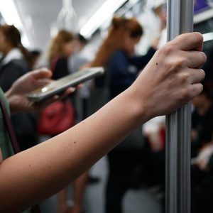 A hand grips a pole while another hand holds a phone on a crowded subway train.