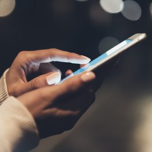 Hands hold a smartphone, tapping the screen, in a dimly lit environment with soft, blurred lights in the background.