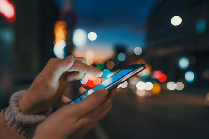 Hand with a smart phone in a dark. Female using her mobile phone, city skyline night light background. Girl pointing finger on screen smart phone on background illumination color light in night city
