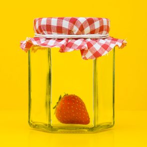 A strawberry sits inside a glass jar, covered with a red-and-white checkered cloth, against a bright yellow background.