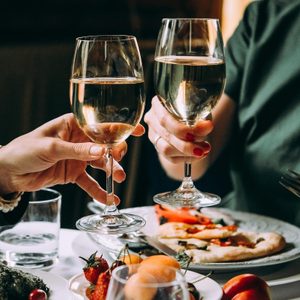 Two wine glasses clink over a table full of dishes, including pizza and fruit, in a cozy dining setting.