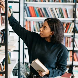 Person reaches for book on a library shelf, surrounded by rows of colorful books.