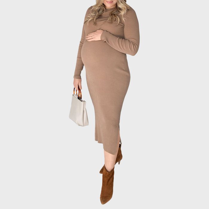 Pregnant woman cradles her belly, wearing a brown dress and boots, holding a white handbag against a plain background.