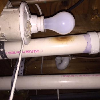Light bulb hangs from ceiling, illuminating PVC pipes marked "Crestline," under wooden floor, with attached pull chain.