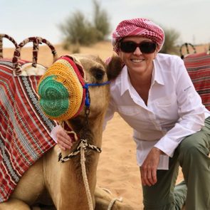 A camel sits in desert sand, wearing a decorative covering. A person in sunglasses and a headscarf kneels beside it, smiling and touching.