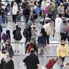 Travelers stand in airport security line, interacting with kiosks and staff. Numerous people carry luggage, wearing masks, within a crowded terminal setting.
