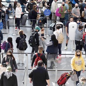 Travelers stand in airport security line, interacting with kiosks and staff. Numerous people carry luggage, wearing masks, within a crowded terminal setting.