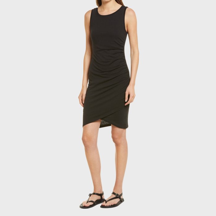 A woman in a sleeveless black dress stands on a plain white background, wearing black sandals.