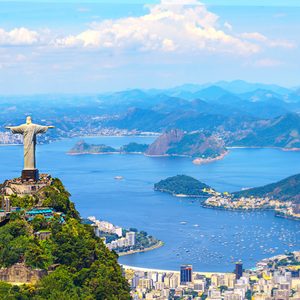 Statue with outstretched arms overlooks city, bay, and mountains; surrounded by lush greenery and buildings.