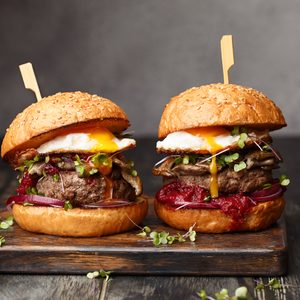 Two burgers topped with sunny-side-up eggs sit on a wooden board, surrounded by a rustic table setting.