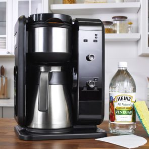 Coffee maker sits idle on a kitchen counter beside a vinegar bottle, sponge, and paper filter, with cabinets and kitchen items in the background.