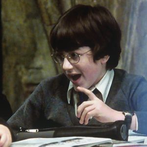 Child wearing glasses talks animatedly, pointing at a table in a dimly lit room.