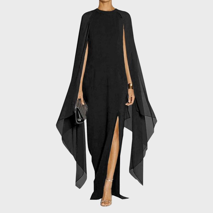 Woman wearing a long black dress with flowing cape sleeves, holding a clutch purse, standing against a plain light gray background.