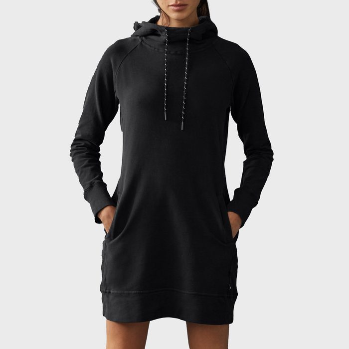 A person wears a black hoodie dress with hands in pockets, standing against a plain gray background.