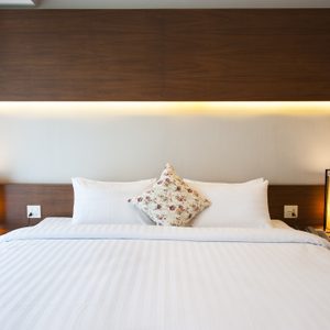 Bed with white sheets and floral pillow, illuminated by modern lamps, set in a room with dark wood accents and light curtains.