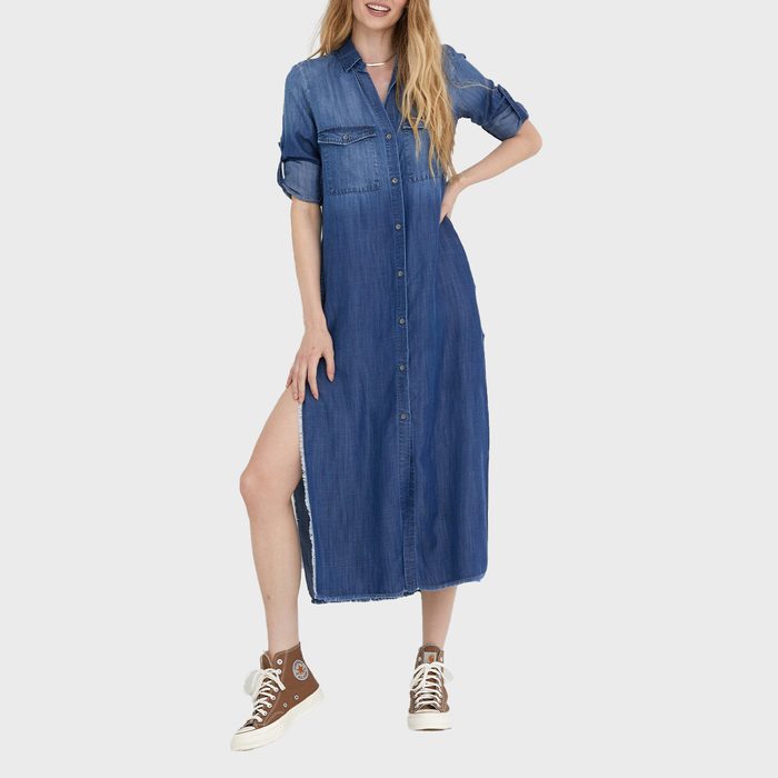 Woman wearing a long denim dress stands confidently, pairing it with brown high-top sneakers, against a plain white background.