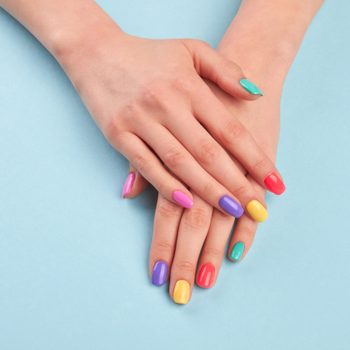 Hands rest on a light blue surface, displaying vibrantly colored nails in pink, purple, yellow, red, and teal.
