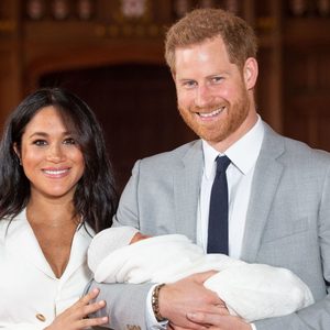 A couple stands smiling, holding a wrapped baby, inside a warmly lit wooden hall.