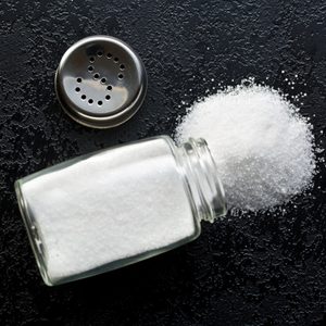 A salt shaker lies on a textured black surface, spilling granular salt, with its metal cap beside it.