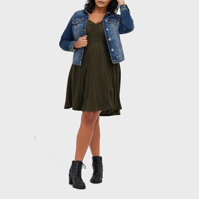 Woman stands wearing a blue denim jacket, olive dress, and black ankle boots, posed against a plain light gray background.