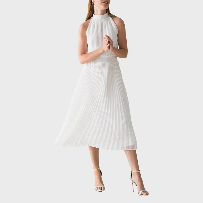 A person wearing a sleeveless, pleated white dress stands, clasping hands with slightly bent knees, against a plain gray background.