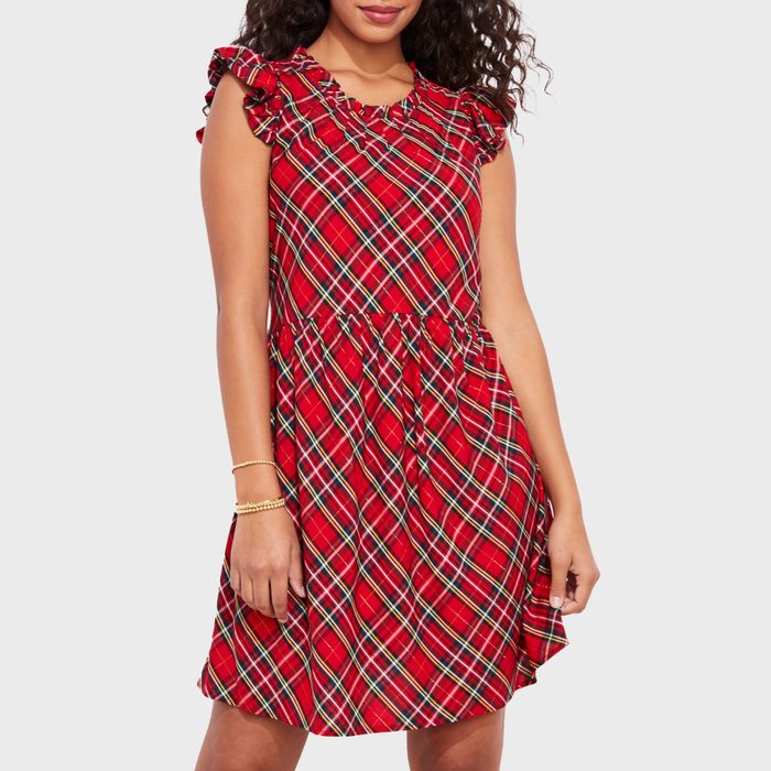 A person wears a sleeveless, red plaid dress with ruffled shoulders, standing casually. The background is plain white.