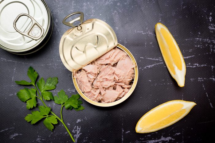 Opened tuna can with parsley and lemon slices on a black cutting board.