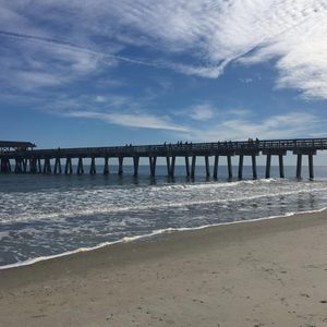 Pier stretches over calm ocean waves, connecting sandy beach to open water under a partly cloudy blue sky.