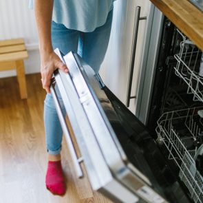 A person opens a dishwasher in a kitchen, with wooden floors and light blue jeans visible.