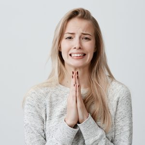 Person pleading with hands clasped, expressing hope or desperation; wearing a gray sweater against a plain, light background.