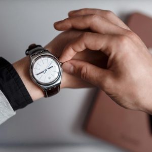 Wristwatch being adjusted by hand, set against a blurred background with a gray sleeve visible on the arm.