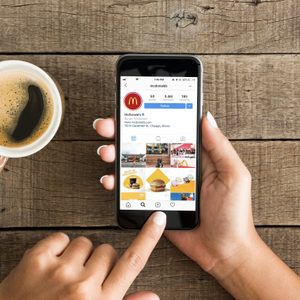 Hand holding smartphone browsing Instagram profile; wooden table surface; coffee cup nearby.