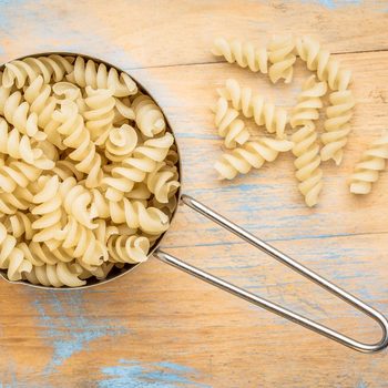 A measuring cup holds fusilli pasta; several pieces spill onto a rustic, blue and orange wooden surface.