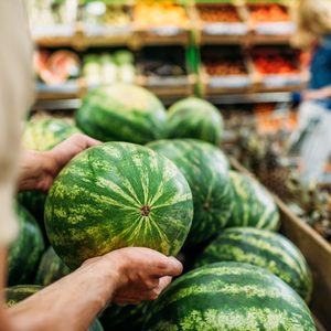 A person selects a watermelon among a pile in a supermarket produce section, with shoppers and fruits in the background.