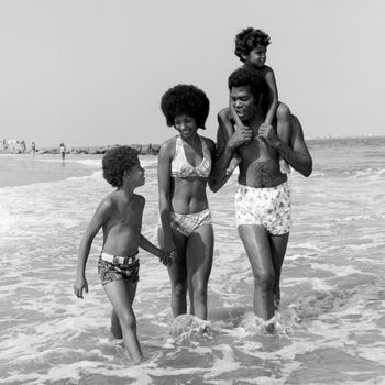 Family walks through shallow ocean waves on a beach, holding hands, enjoying the sunny day together.