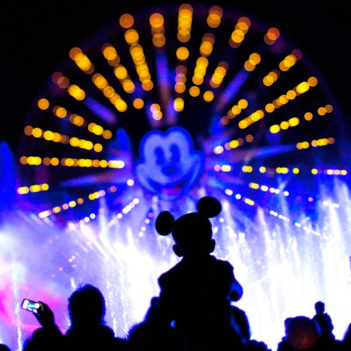 Silhouette of mouse ears worn by person lit against vibrant, colorful water and light show, featuring a large illuminated wheel with a central cartoon face.