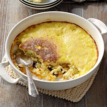 A baked pudding sits in a round dish, partially scooped with a spoon, resting on a knitted mat on a wooden surface.