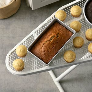 Loaf of bread cooling on an ironing board with muffins, surrounded by a light kitchen environment.