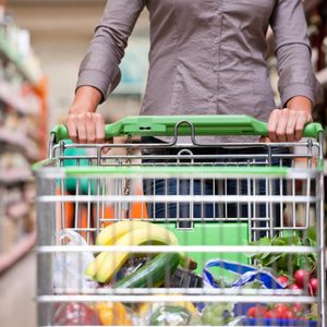 A person pushes a shopping cart loaded with groceries, including bananas, through a supermarket aisle lined with shelves.