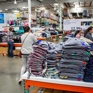 Stacks of clothes lie on a table while shoppers browse in a large warehouse. Signs display prices, including 