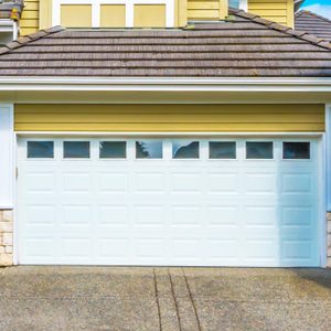 A closed white garage door with windows, surrounded by a yellow house exterior and green shrubs.